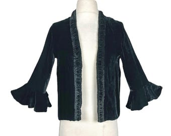 May include: A dark teal velvet jacket with ruffled trim and flared sleeves. The open-front jacket is displayed on a mannequin against a white background. The sleeves have a bell-like shape.
