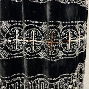 May include: Close-up of a black velvet garment with intricate silver beadwork. The design features geometric patterns, including crosses and repeating shapes, creating a luxurious and detailed texture. The beadwork is meticulously applied, adding a shimmering effect.