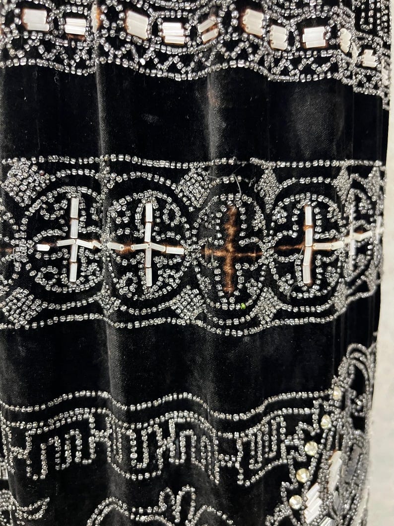 May include: Close-up of a black velvet garment with intricate silver beadwork. The design features geometric patterns, including crosses and repeating shapes, creating a luxurious and detailed texture. The beadwork is meticulously applied, adding a shimmering effect.