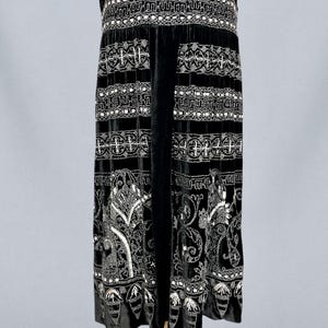 May include: A black velvet dress with intricate white beaded embellishments. The dress features a detailed pattern around the waist and along the skirt, with a decorative hemline. The sleeveless design and elegant detailing suggest a vintage or formal style.