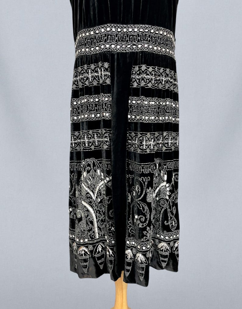 May include: A black velvet dress with intricate white beaded embellishments. The dress features a detailed pattern around the waist and along the skirt, with a decorative hemline. The sleeveless design and elegant detailing suggest a vintage or formal style.