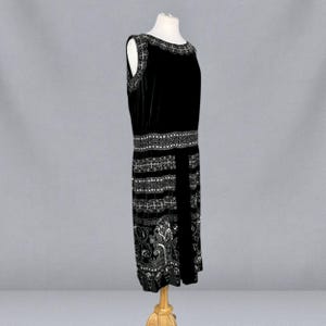 May include: A black sleeveless dress with intricate white embellishments. The dress features a detailed pattern around the neckline, waist, and hem. The design includes floral and geometric elements, creating a vintage aesthetic. The dress is displayed on a mannequin.
