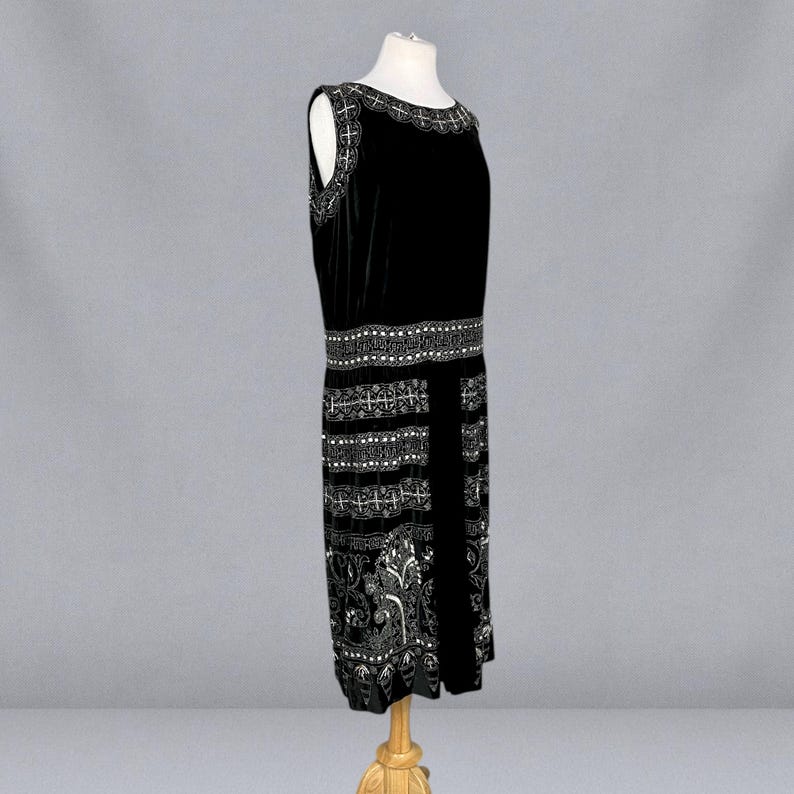 May include: A black sleeveless dress with intricate white embellishments. The dress features a detailed pattern around the neckline, waist, and hem. The design includes floral and geometric elements, creating a vintage aesthetic. The dress is displayed on a mannequin.