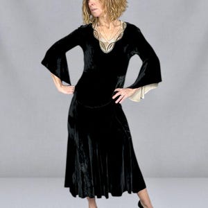 Puede incluir: Un vestido de terciopelo negro con mangas largas y acampanadas y una falda midi. El escote est&aacute; adornado con bordados dorados y crema. El vestido se combina con tacones negros, creando un look de inspiraci&oacute;n vintage.