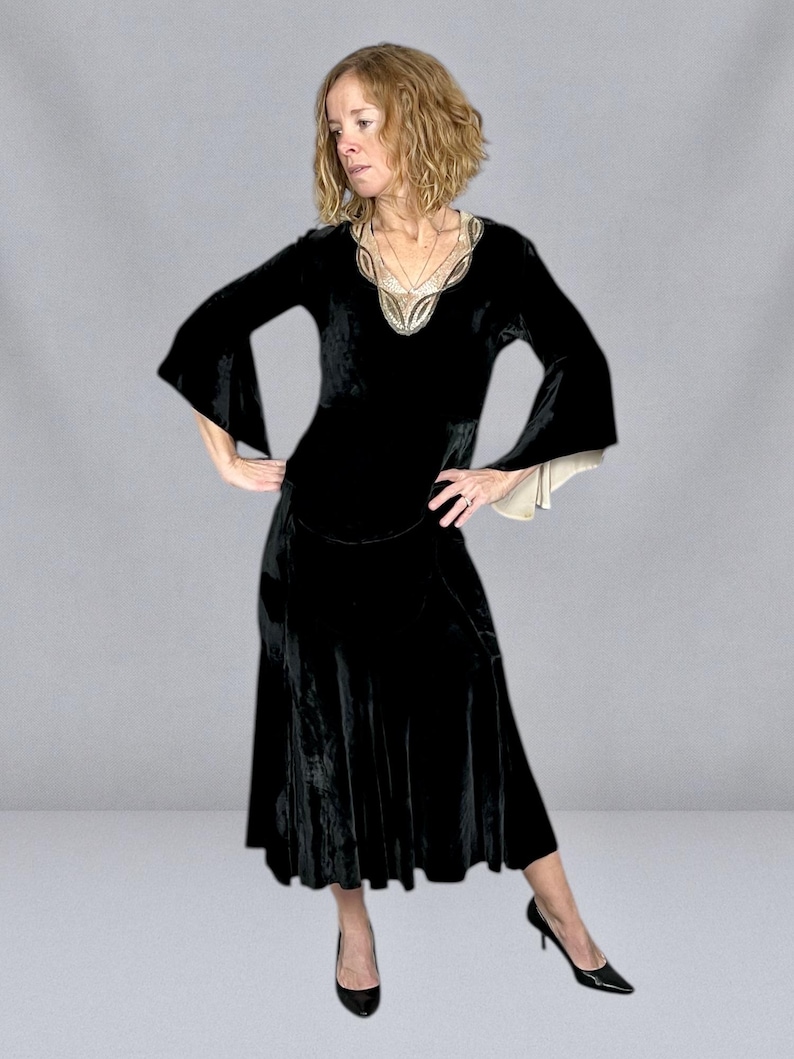 Puede incluir: Un vestido de terciopelo negro con mangas largas y acampanadas y una falda midi. El escote est&aacute; adornado con bordados dorados y crema. El vestido se combina con tacones negros, creando un look de inspiraci&oacute;n vintage.