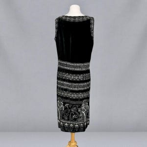 May include: A sleeveless black velvet dress with intricate silver-colored embroidery. The dress features a detailed pattern around the neckline, waist, and hem, with a floral design at the bottom. The dress is displayed on a mannequin.