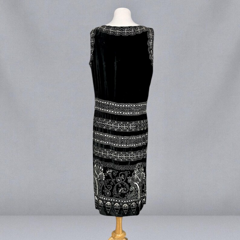 May include: A sleeveless black velvet dress with intricate silver-colored embroidery. The dress features a detailed pattern around the neckline, waist, and hem, with a floral design at the bottom. The dress is displayed on a mannequin.