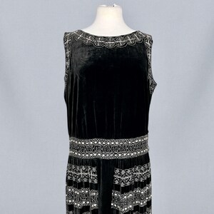 May include: A sleeveless black velvet dress with intricate silver beaded embellishments. The dress features a detailed beaded neckline, a decorative belt, and a patterned hemline. The dress is displayed on a mannequin.