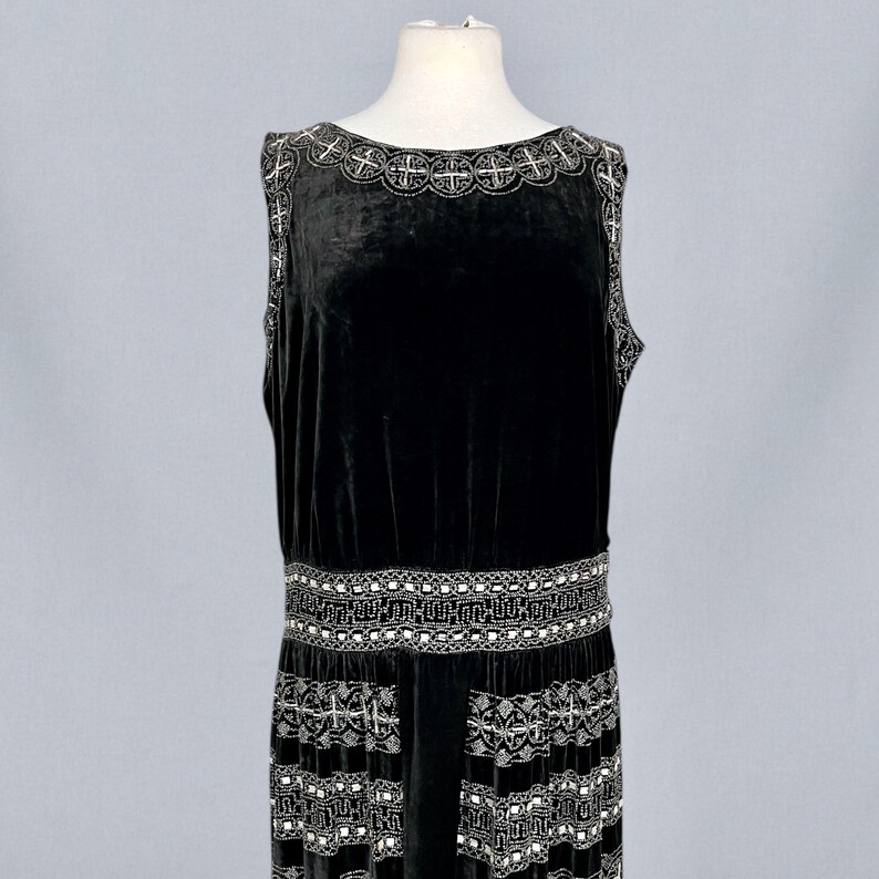 May include: A sleeveless black velvet dress with intricate silver beaded embellishments. The dress features a detailed beaded neckline, a decorative belt, and a patterned hemline. The dress is displayed on a mannequin.