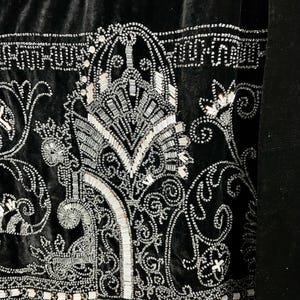 May include: Close-up of a black velvet textile with intricate silver beadwork. The design features ornate floral and geometric patterns, with a central stylized motif. The beadwork creates a shimmering effect against the dark background.