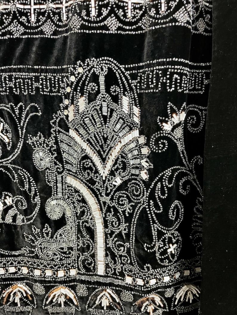 May include: Close-up of a black velvet textile with intricate silver beadwork. The design features ornate floral and geometric patterns, with a central stylized motif. The beadwork creates a shimmering effect against the dark background.