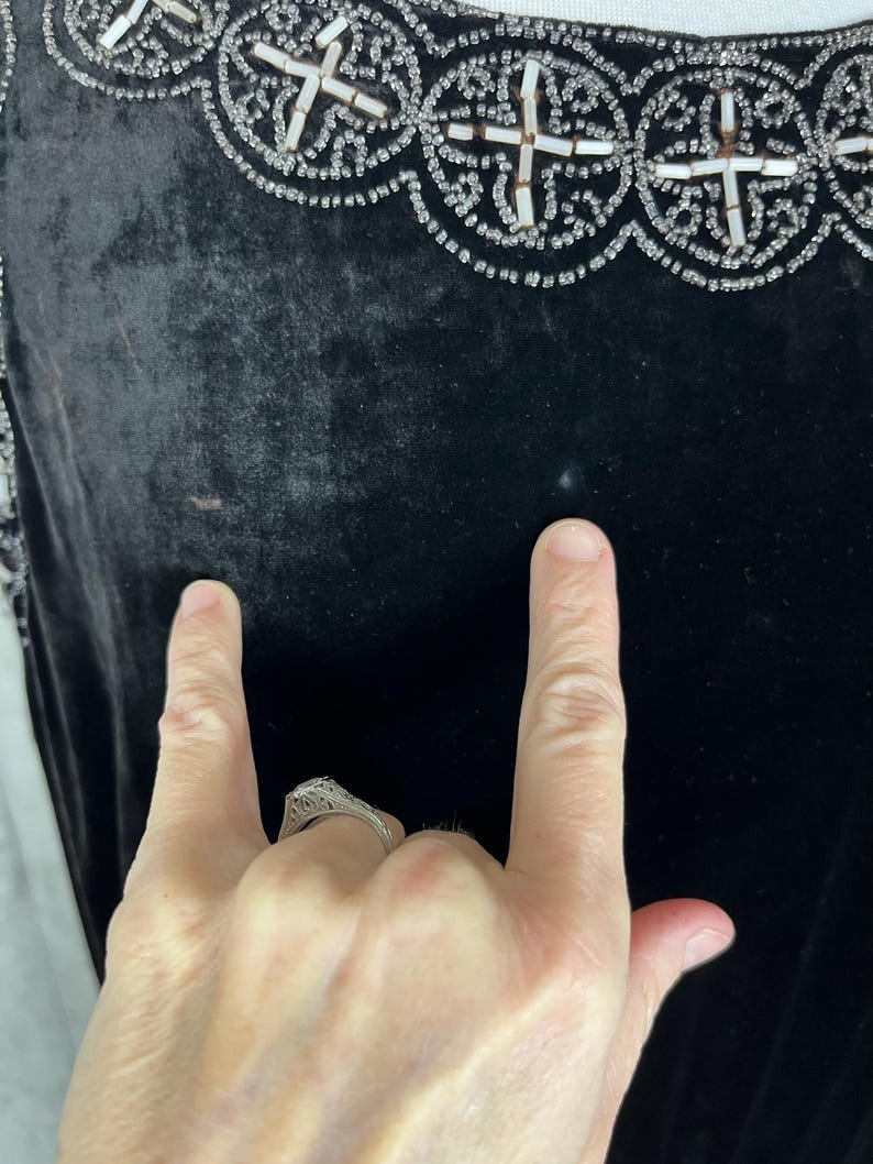 May include: Close-up of a black velvet garment with ornate beaded embellishments. The trim features a repeating pattern of white and silver beads. The garment's fabric appears soft and luxurious, with a slight sheen. The image shows a portion of the garment.