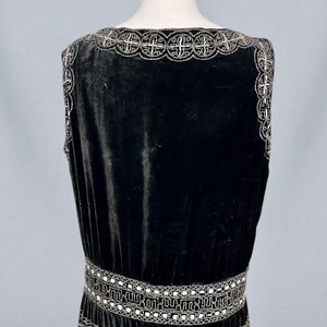May include: A black velvet dress with silver beaded embellishments. The dress features a square neckline and a detailed beaded design around the neckline, waist, and hem. The dress is displayed on a mannequin.