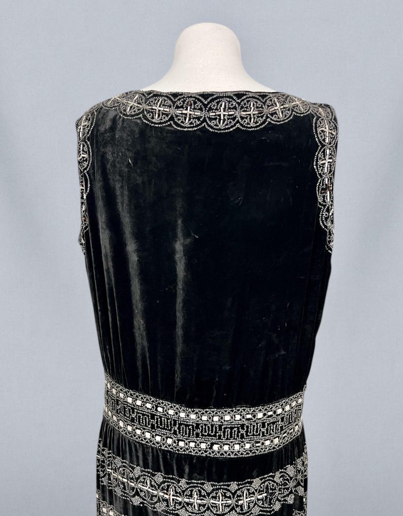 May include: A black velvet dress with silver beaded embellishments. The dress features a square neckline and a detailed beaded design around the neckline, waist, and hem. The dress is displayed on a mannequin.