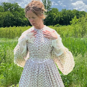 May include: A woman wearing a white dress with a floral pattern and lace trim. The dress has a fitted bodice and a full skirt. She is standing in a field of tall grass.