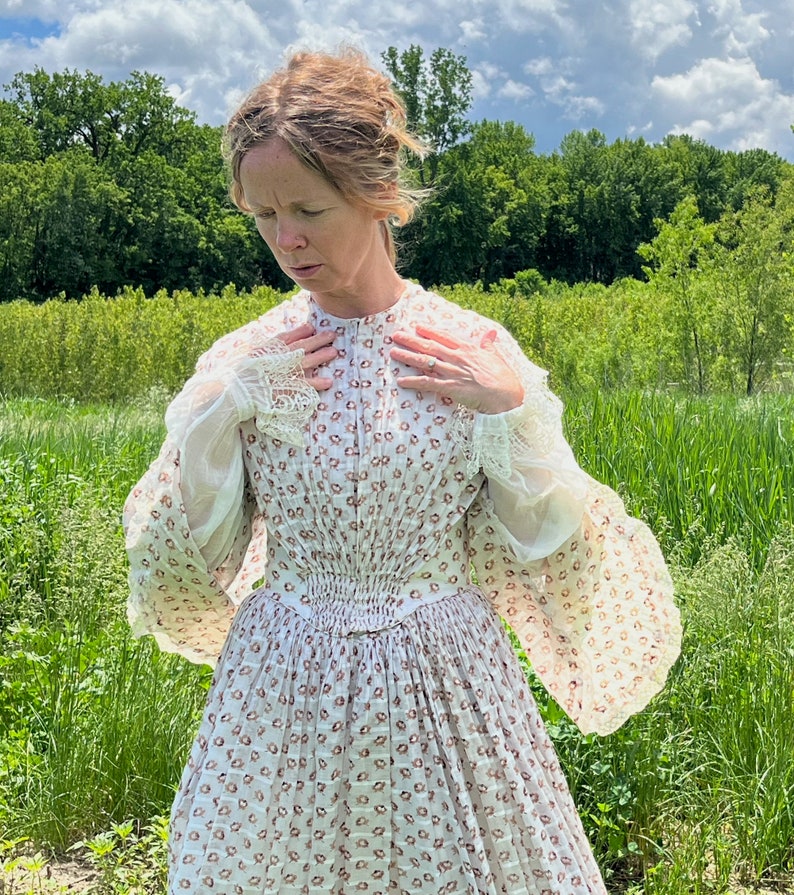 May include: A woman wearing a white dress with a floral pattern and lace trim. The dress has a fitted bodice and a full skirt. She is standing in a field of tall grass.