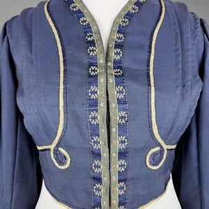 May include: A navy blue jacket with intricate trim. The jacket features a V-neck design with decorative trim along the edges and a detailed pattern on the front. The sleeves are long and the jacket has a cropped fit.