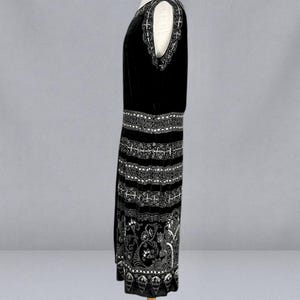 May include: A black sleeveless dress with intricate white embroidery. The dress features a detailed pattern around the neckline, waist, and hem, with a floral design on the lower portion. The dress is displayed on a mannequin.