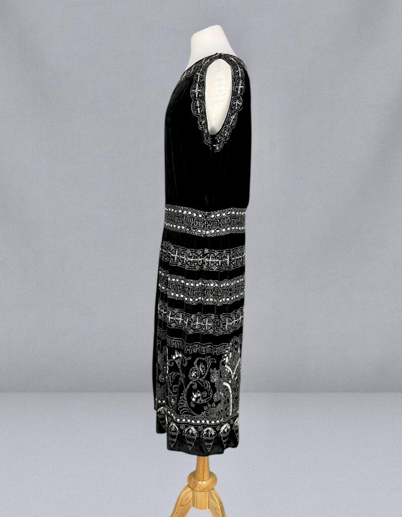 May include: A black sleeveless dress with intricate white embroidery. The dress features a detailed pattern around the neckline, waist, and hem, with a floral design on the lower portion. The dress is displayed on a mannequin.