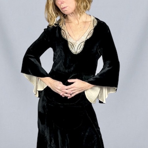 Puede incluir: Vestido de terciopelo negro con escote en V y mangas largas acampanadas. El escote est&aacute; adornado con un dise&ntilde;o de cuentas en tonos crema y plata. Las mangas tienen un forro color crema que se asoma en los pu&ntilde;os. El vestido parece vintage.