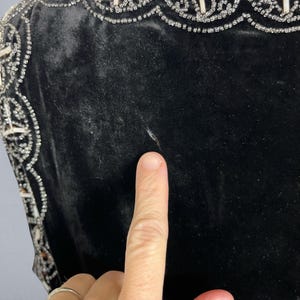 May include: Close-up of a black velvet garment with ornate, beaded trim. The trim features a repeating pattern of white beads and clear crystals. The fabric appears soft and luxurious. A finger points to the garment.