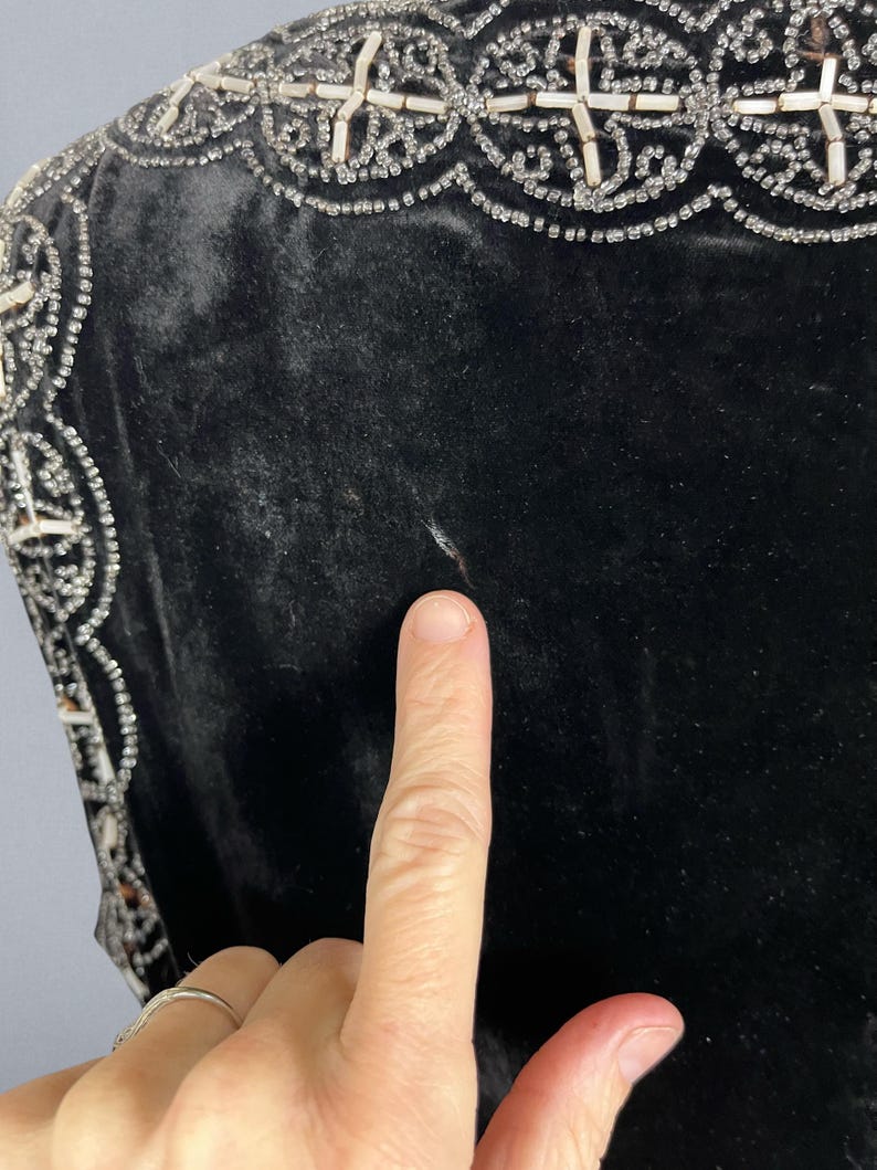 May include: Close-up of a black velvet garment with ornate, beaded trim. The trim features a repeating pattern of white beads and clear crystals. The fabric appears soft and luxurious. A finger points to the garment.
