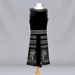 May include: A sleeveless black velvet dress with white embroidered detailing. The dress features a decorative neckline, a detailed waist band, and a patterned skirt. The dress is displayed on a mannequin.