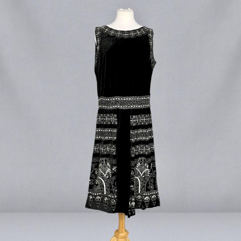 May include: A sleeveless black velvet dress with white embroidered detailing. The dress features a decorative neckline, a detailed waist band, and a patterned skirt. The dress is displayed on a mannequin.