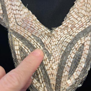 Puede incluir: Primer plano de un cuello con cuentas vintage. El cuello presenta un dise&ntilde;o de cuello en V con una delicada base de malla blanquecina. El cuello est&aacute; adornado con hileras de peque&ntilde;as cuentas rectangulares en tonos crema, plata y rosa p&aacute;lido.