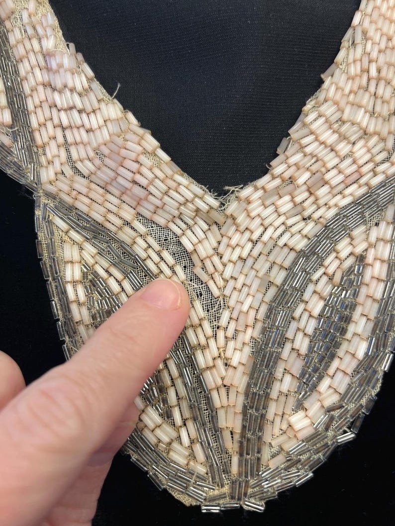 Puede incluir: Primer plano de un cuello con cuentas vintage. El cuello presenta un dise&ntilde;o de cuello en V con una delicada base de malla blanquecina. El cuello est&aacute; adornado con hileras de peque&ntilde;as cuentas rectangulares en tonos crema, plata y rosa p&aacute;lido.