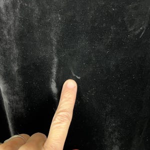 May include: Close-up of black velvet fabric. The fabric has a soft, textured surface with subtle variations in tone. A finger is pointing at the fabric, highlighting its texture and color. The velvet appears to be in good condition.