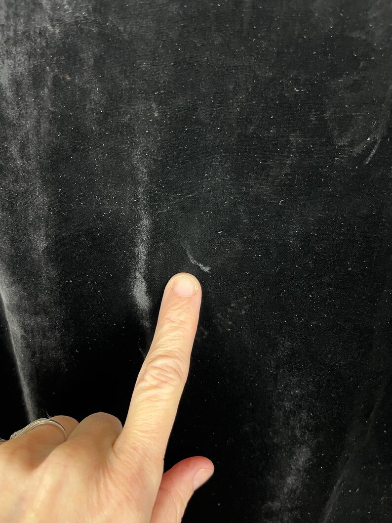 May include: Close-up of black velvet fabric. The fabric has a soft, textured surface with subtle variations in tone. A finger is pointing at the fabric, highlighting its texture and color. The velvet appears to be in good condition.