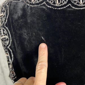 May include: Close-up of a black velvet garment with ornate beaded embellishments. The trim features a repeating pattern of white beads and silver accents. The fabric appears soft and luxurious.