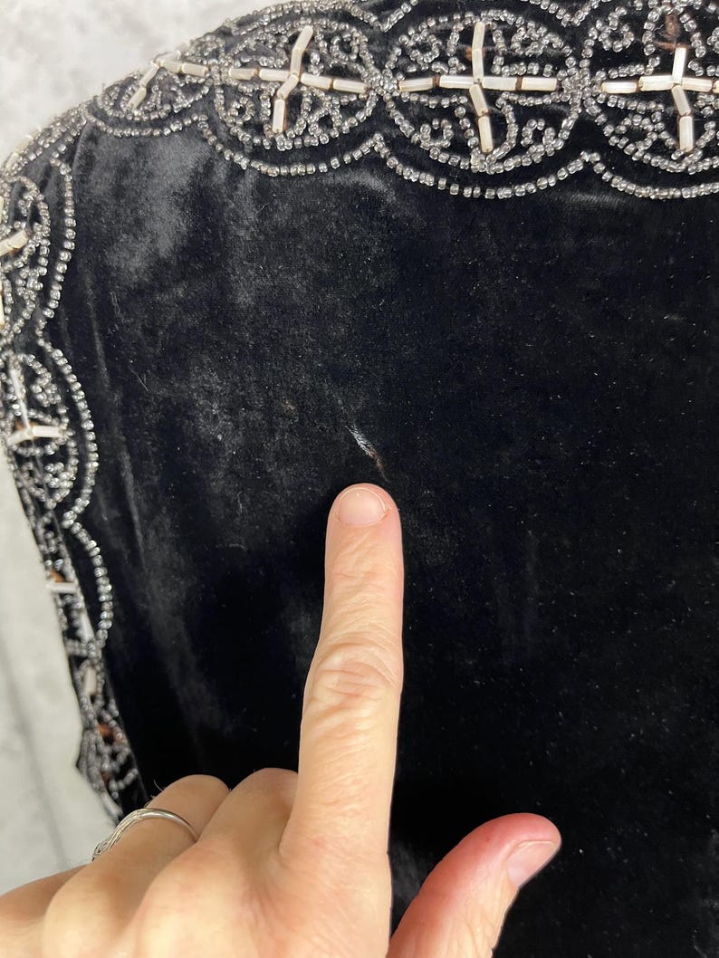 May include: Close-up of a black velvet garment with ornate beaded embellishments. The trim features a repeating pattern of white beads and silver accents. The fabric appears soft and luxurious.