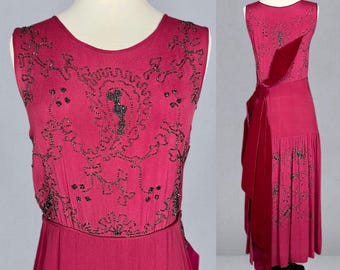 May include: A sleeveless, magenta-colored dress with intricate black beadwork on the bodice. The dress features a flowing skirt and a long, draped sash. The dress is displayed on a mannequin, showcasing its vintage style and elegant design.