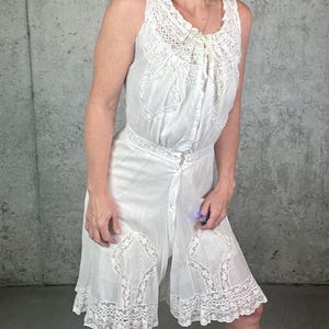 May include: A white sleeveless romper featuring intricate lace embroidery and cutwork detailing along the neckline, waist, and hem. The garment includes a front button closure and a drawstring tie at the collar, with a ruffled lace trim at the leg openings.