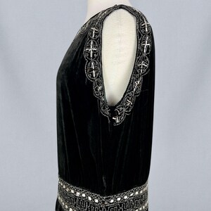 May include: A black velvet dress with intricate silver beaded embellishments. The dress features a scoop neckline and a drop waist, with detailed beading along the shoulders and hemline. The dress is displayed on a mannequin.