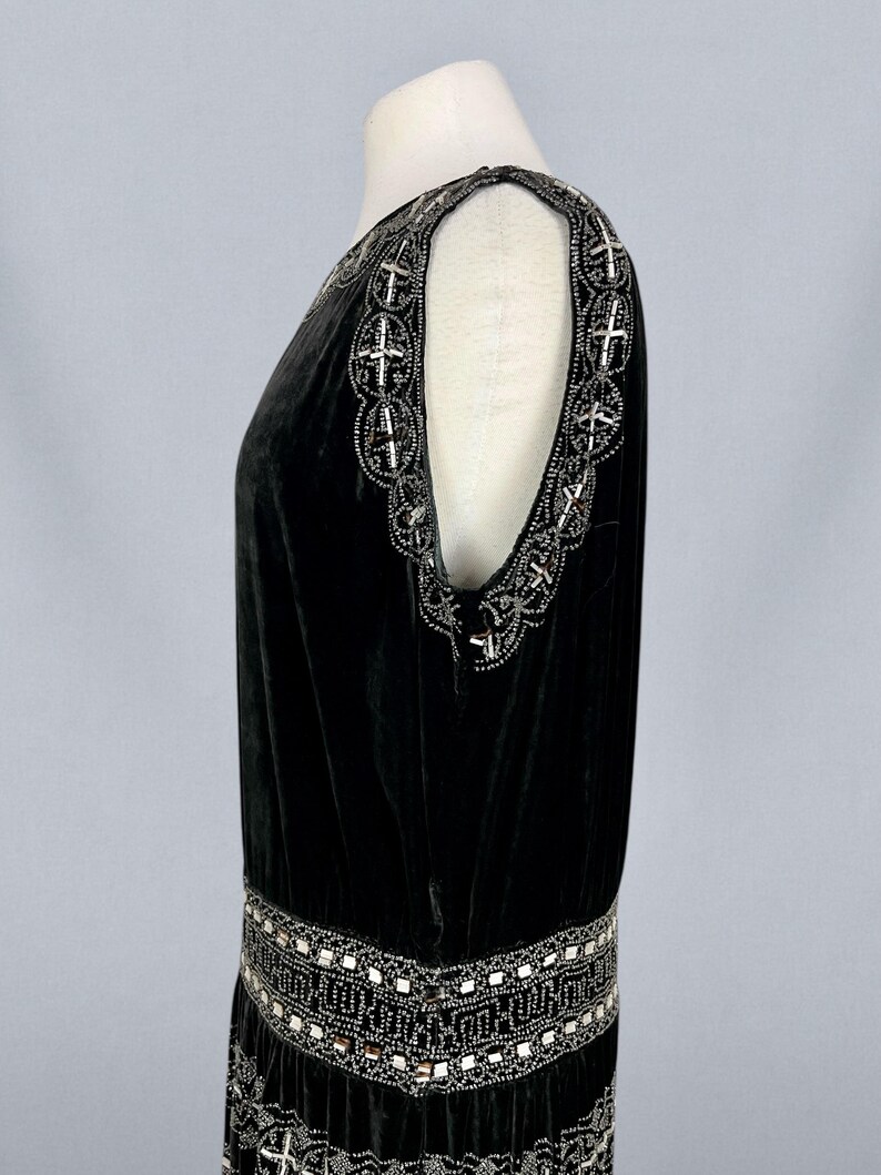 May include: A black velvet dress with intricate silver beaded embellishments. The dress features a scoop neckline and a drop waist, with detailed beading along the shoulders and hemline. The dress is displayed on a mannequin.