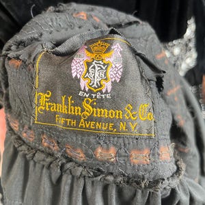 May include: Close-up of a black garment label with the text "Franklin Simon & Co. Fifth Avenue, N.Y." in gold lettering. The label also features a crest with a crown and the words "En T&ecirc;te". The garment has a ruffled edge.