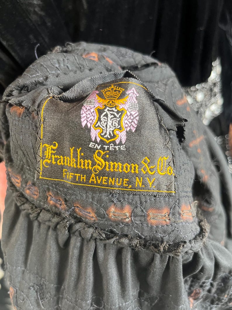 May include: Close-up of a black garment label with the text "Franklin Simon & Co. Fifth Avenue, N.Y." in gold lettering. The label also features a crest with a crown and the words "En T&ecirc;te". The garment has a ruffled edge.