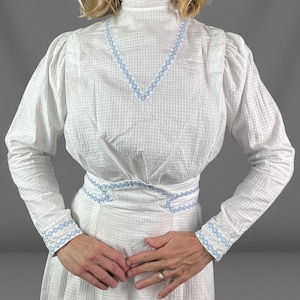 May include: A white long-sleeved blouse featuring a subtle checkered texture and a high collar. The garment is accented with light blue geometric embroidery along the neckline, waist, and cuffs, creating a structured look with puff sleeves.