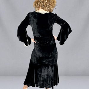 Puede incluir: Un vestido de terciopelo negro con mangas largas y acampanadas y una falda escalonada. El vestido tiene un cuello decorativo y se combina con tacones negros. El modelo se ve de espaldas.