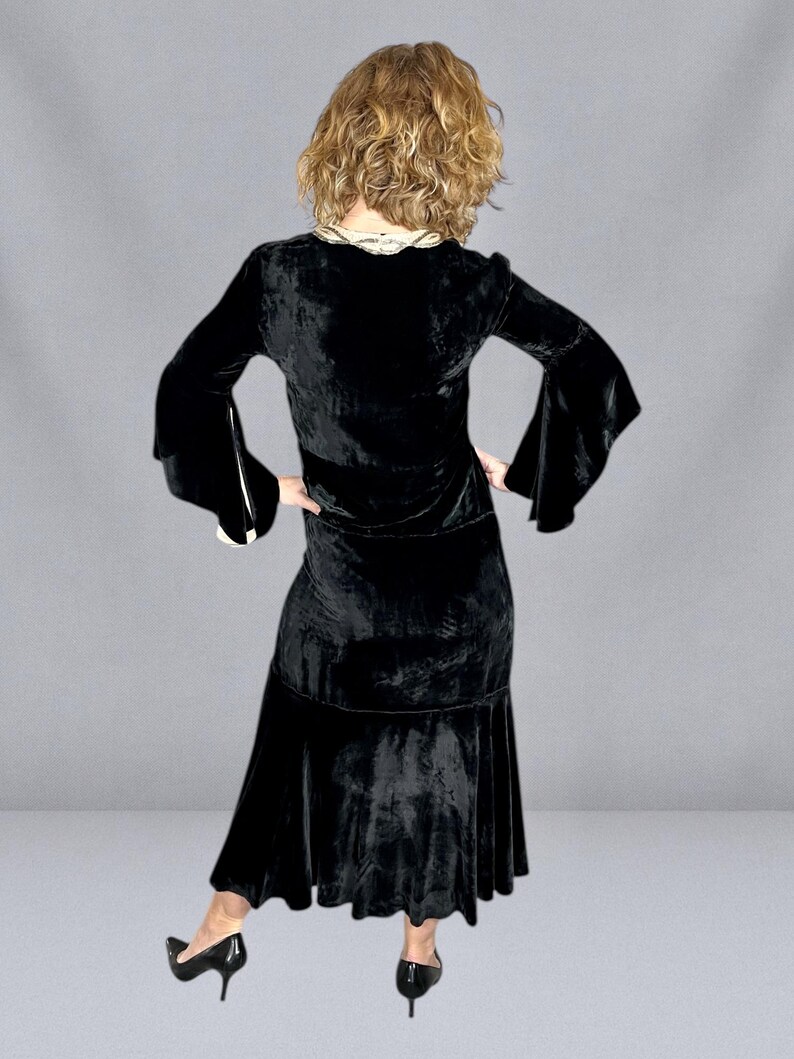 Puede incluir: Un vestido de terciopelo negro con mangas largas y acampanadas y una falda escalonada. El vestido tiene un cuello decorativo y se combina con tacones negros. El modelo se ve de espaldas.