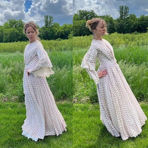 May include: A white dress with a floral pattern and lace trim. The dress has long sleeves and a high neckline. The dress is photographed outdoors in a field of green grass.