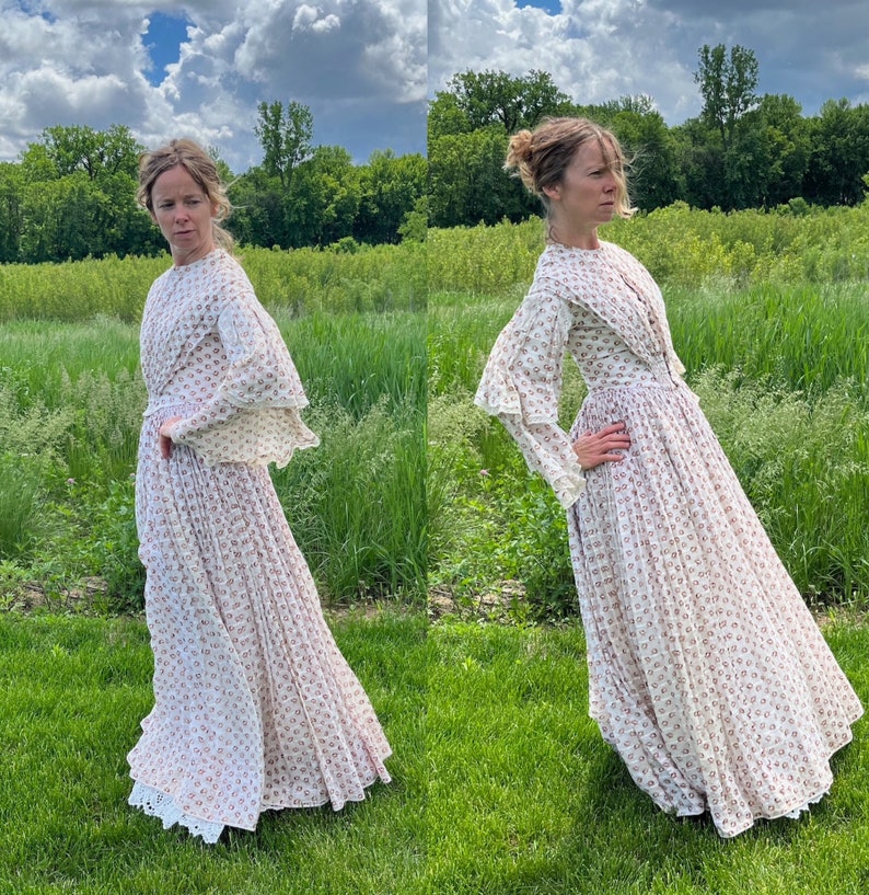 May include: A white dress with a floral pattern and lace trim. The dress has long sleeves and a high neckline. The dress is photographed outdoors in a field of green grass.