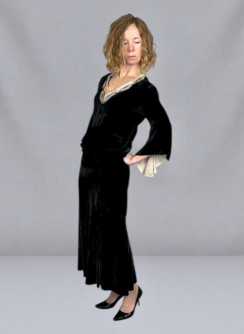Puede incluir: Un vestido largo de terciopelo negro con escote en V y mangas acampanadas. El escote est&aacute; adornado con una tela color crema con detalles dorados. El vestido se combina con tacones negros.