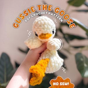 May include: A crocheted white goose with a yellow beak and feet. The text "GUSSIE THE GOOSE PDF CROCHET PATTERN" is displayed in orange. The bottom right corner has a sticker that says "NO SEW!"