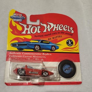 May include: A vintage Hot Wheels toy car on its original packaging. The packaging is red and yellow with the Hot Wheels logo and text. The car is red with black wheels and is in a clear plastic case. Includes a matching collector's button.