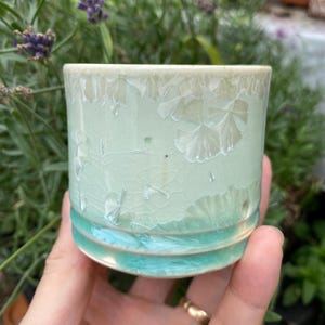 Op de afbeelding: Een kleine, handgemaakte keramische beker met een lichtgroen glazuur en turkooizen banden. De beker heeft een uniek kristallijn patroon op het bovenste deel, wat een gestructureerd, artistiek element toevoegt. De beker wordt in een hand gehouden.