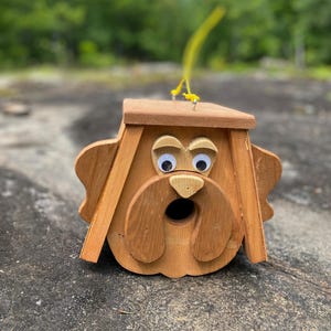May include: A wooden birdhouse shaped like a dog's face. The birdhouse is made of light brown wood with googly eyes and a round opening for the birds. A yellow string is attached to the top for hanging.